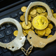 A pair of handcuffs with one cuff open on top of a pile of gold coins bearing the bitcoin logo. This all sits on top of a laptop keyboard.